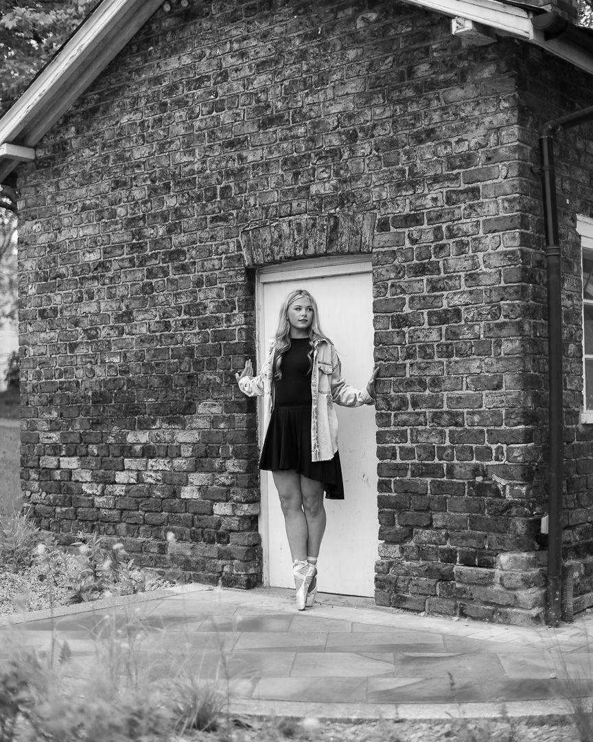 A blonde woman in a black dress and denim jacket poses by a white door of a rustic brick building in black and white.