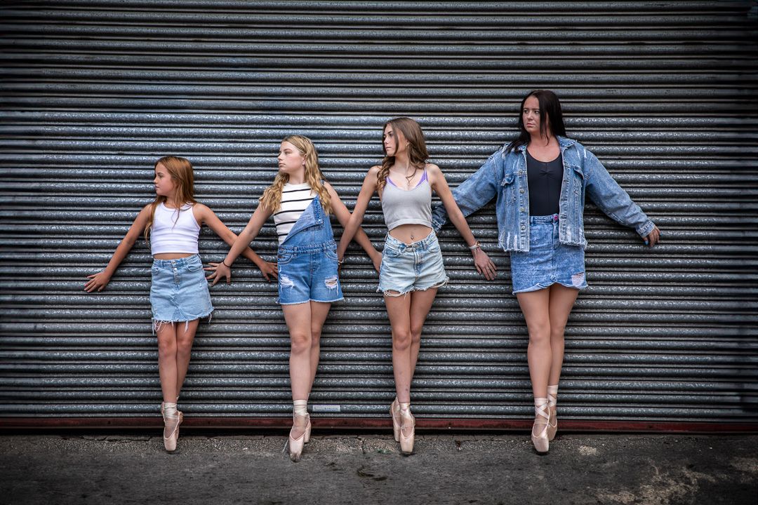 Row of ballet dancers sitting against industrial wall wearing denim and pointe shoes.