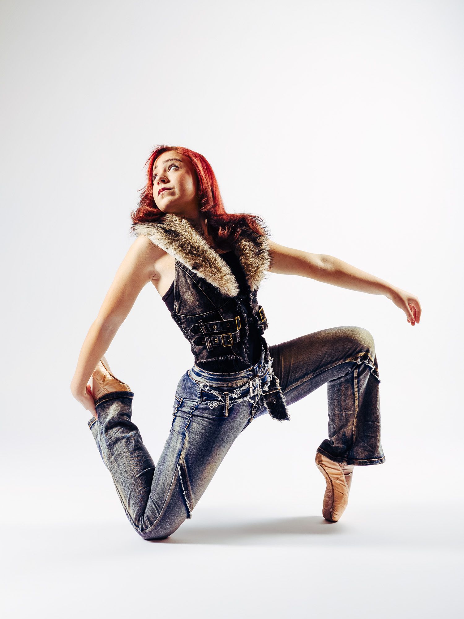 Red-haired female dancer in jeans and fur vest striking a dynamic pose on pointe in a studio.