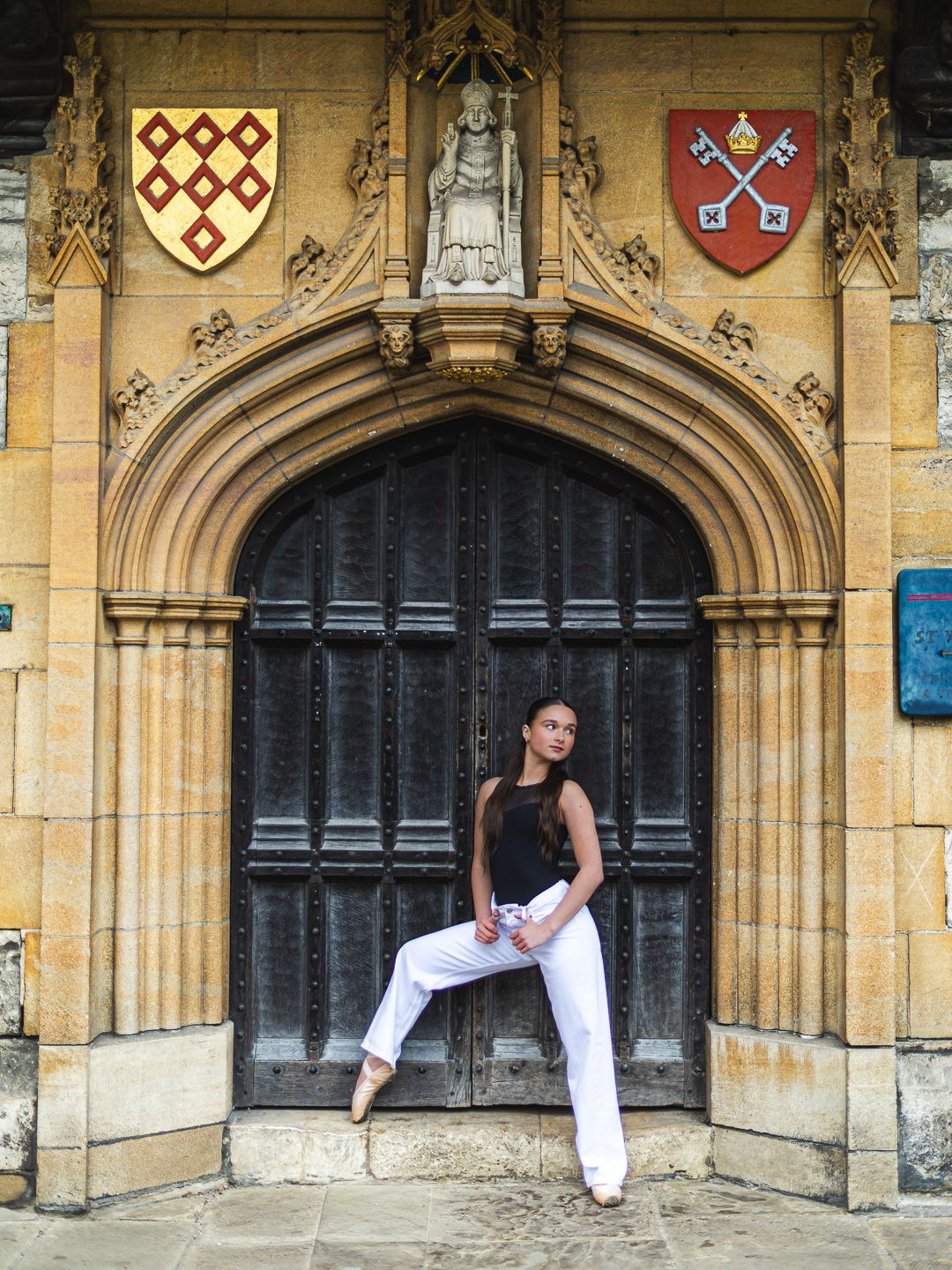Woman in white pants and black top posing before ornate Gothic arched doorway with heraldic crests.