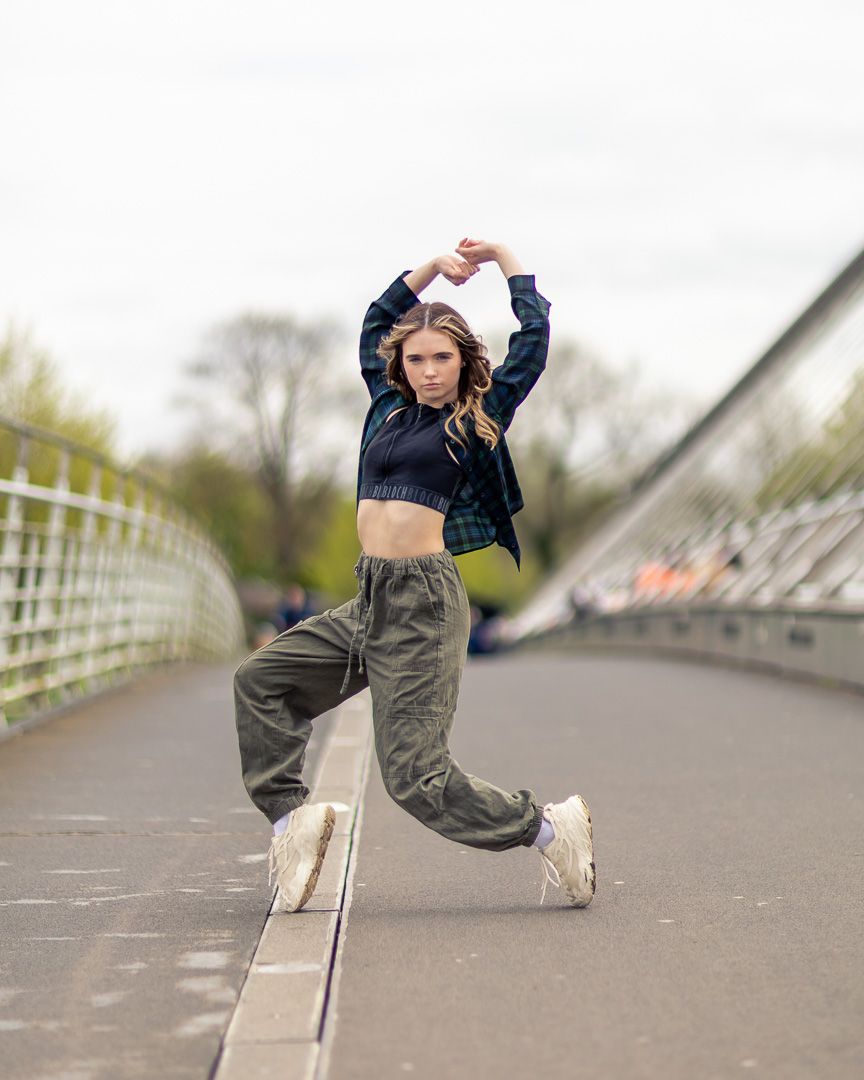 A young woman in cargo pants and crop top jumps mid-air on a bridge, striking a dynamic dance pose outdoors.