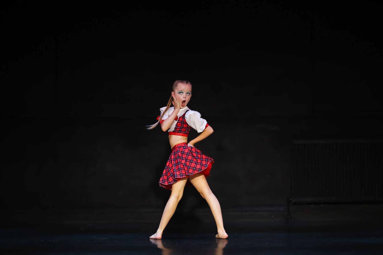 Young dancer performs on stage in a red plaid skirt and white blouse costume under dramatic spotlight.