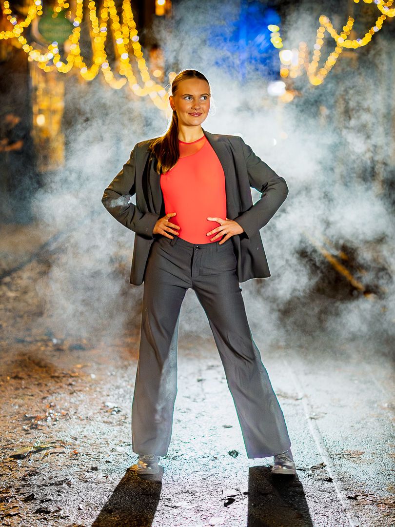 A fashion model poses dramatically in a grey suit and red top against a smoky backdrop with twinkling lights.