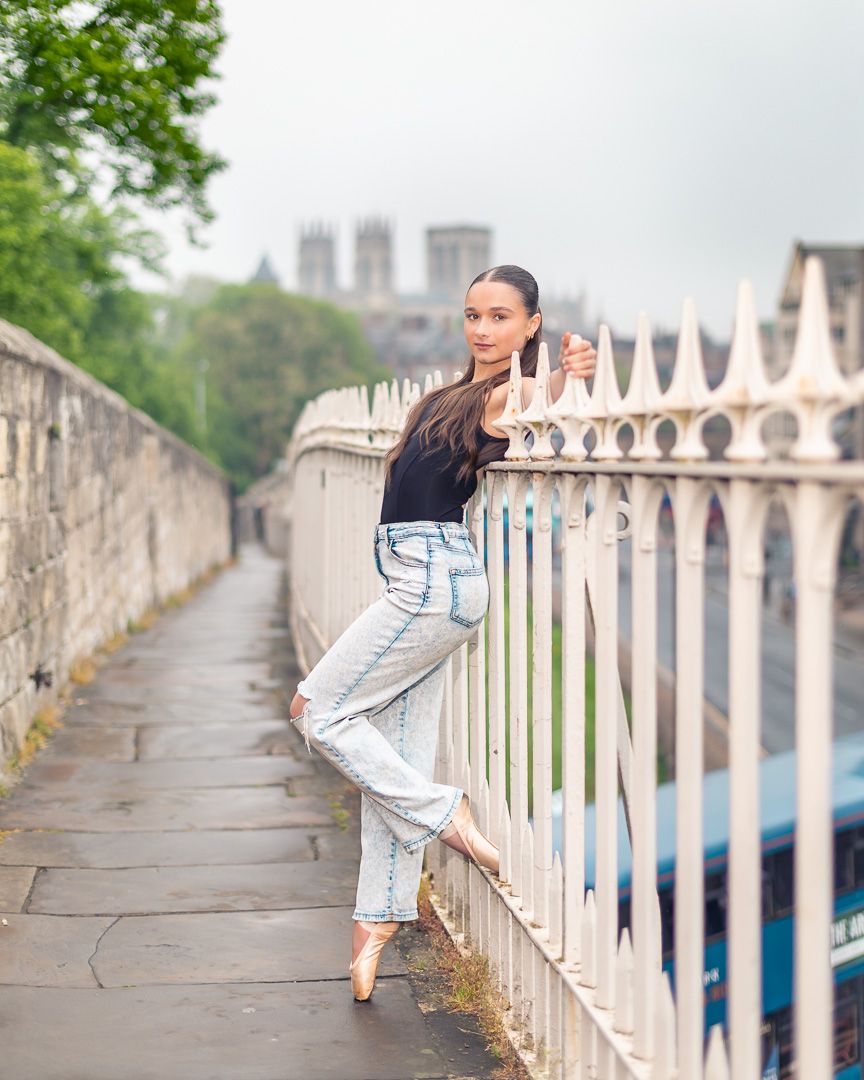 A young woman in jeans and black top poses gracefully on pointe shoes against white iron fence with York Minster in background.