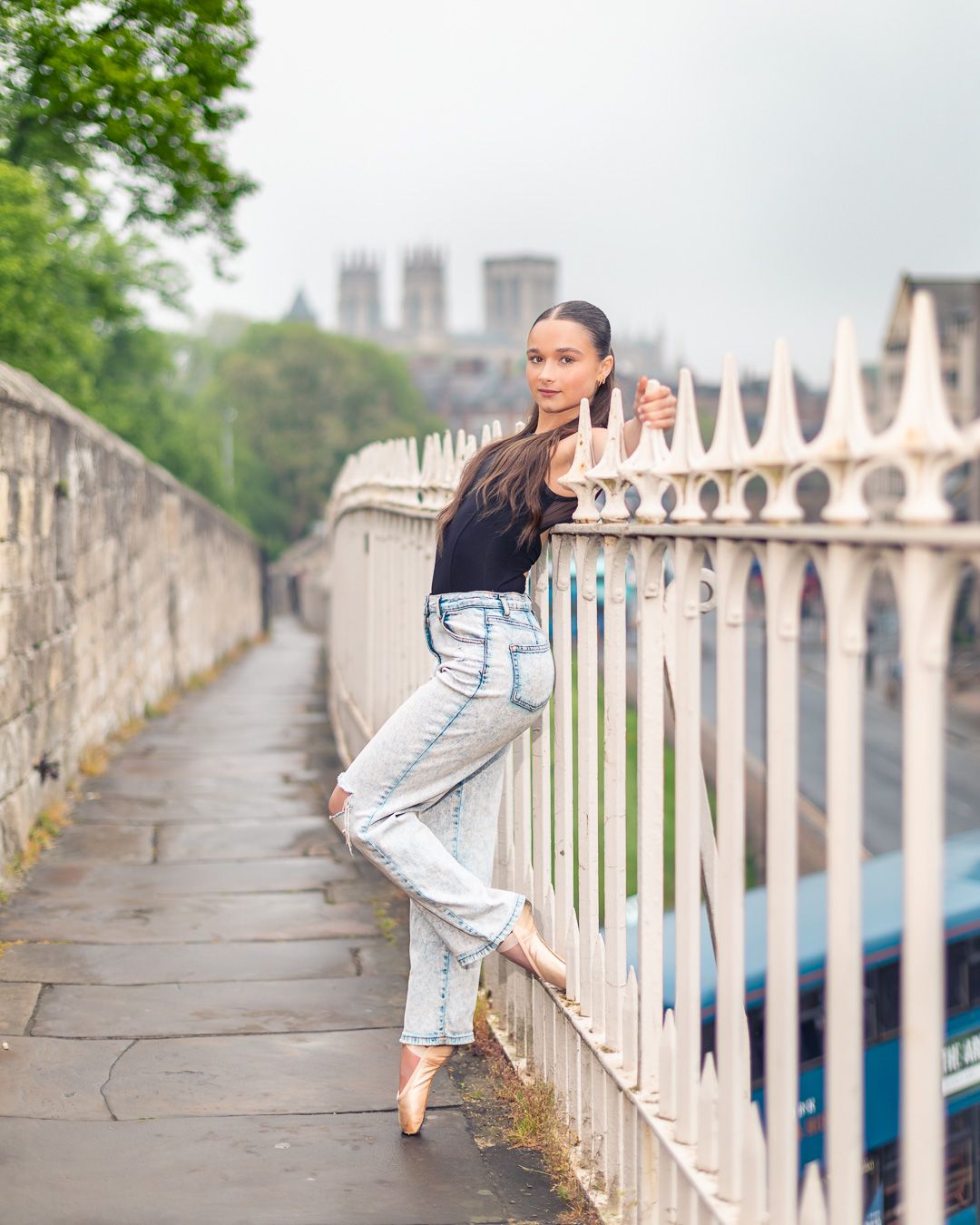 A young woman in jeans and black top poses gracefully on white iron fence along a historic stone pathway.
