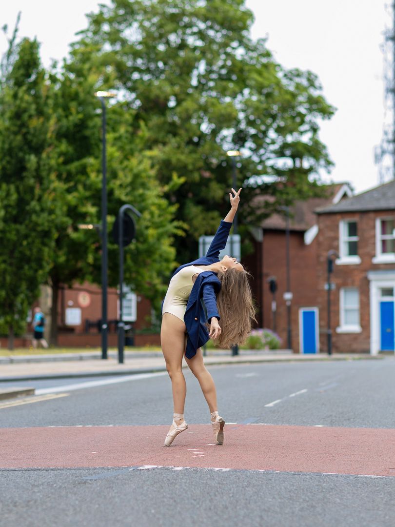A ballet dancer in a blue blazer and leotard performs an arabesque on pointe in the middle of a street.