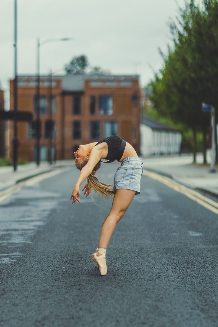 Ballet dancer performs elegant pose on asphalt road with urban buildings in background.