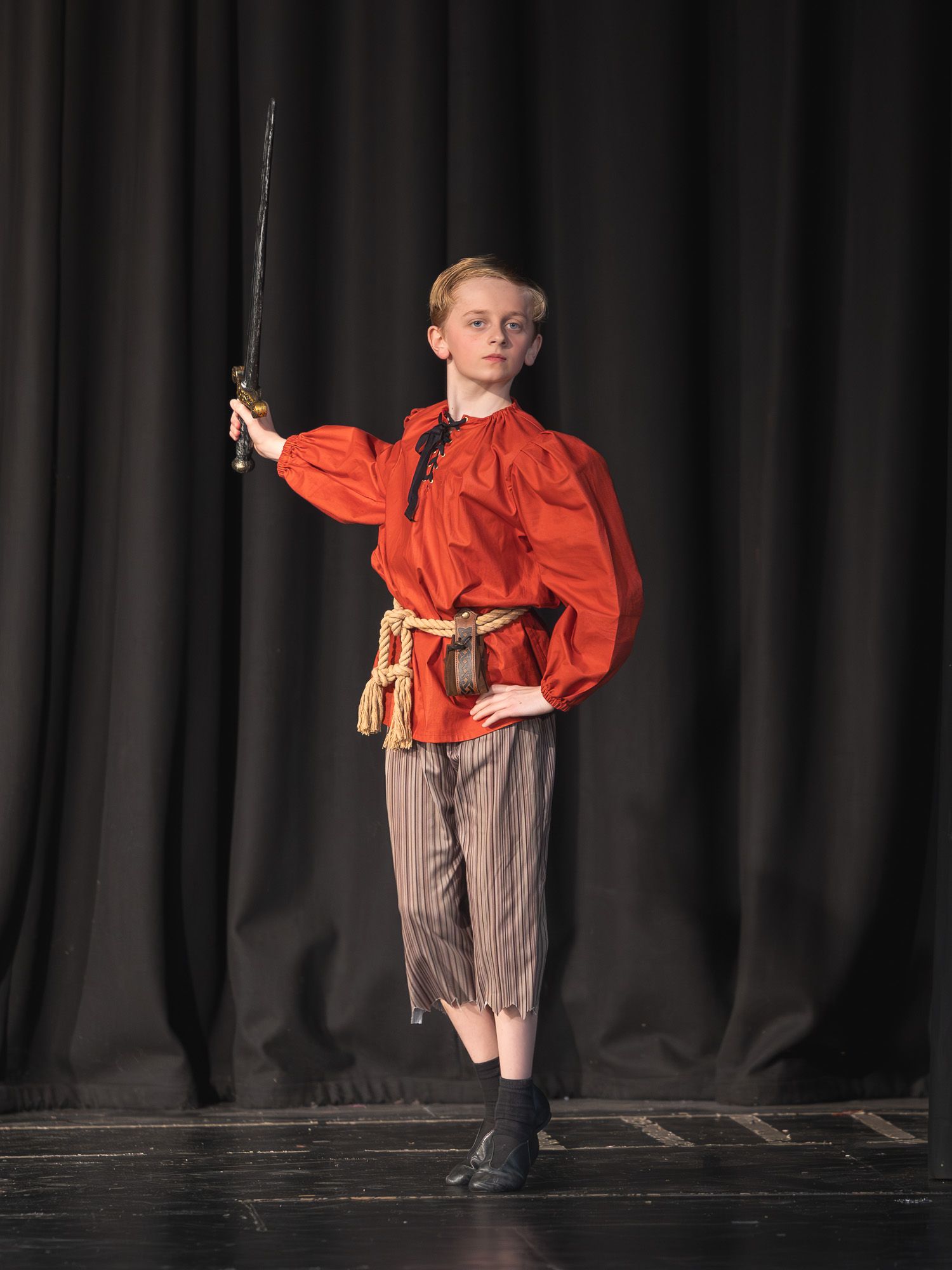 Young performer in red pirate costume holding sword on stage during school theater production.