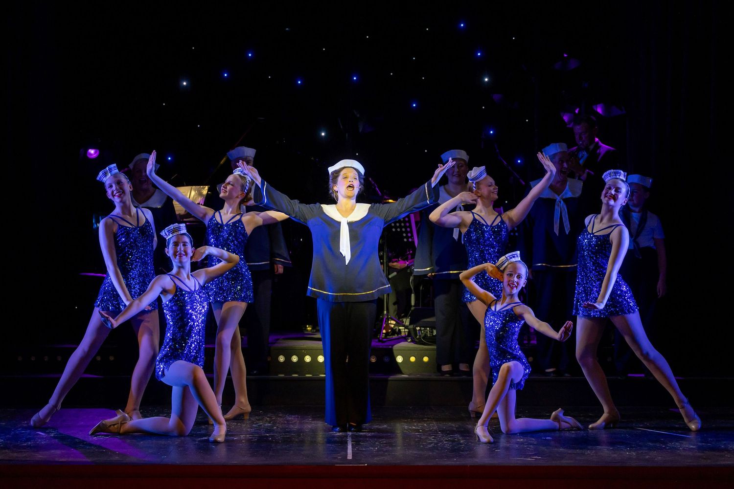 Sailor-costumed performer center stage with dancers in blue sequined outfits during a colorful Broadway musical production number.