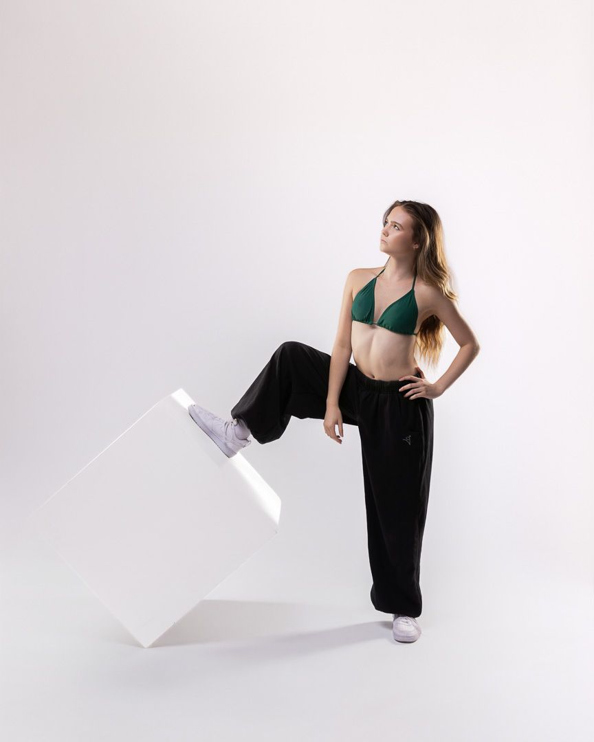 Woman in green bikini top and black wide-leg pants poses confidently beside a white geometric cube prop.