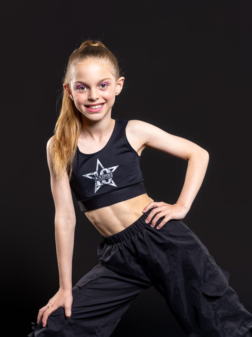 Young dancer in black crop top and baggy pants poses confidently against dark background.