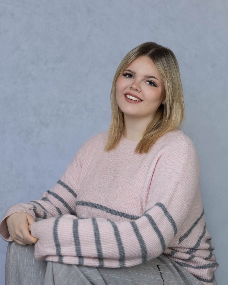 Young woman in pink striped sweater smiling while seated against a gray background.