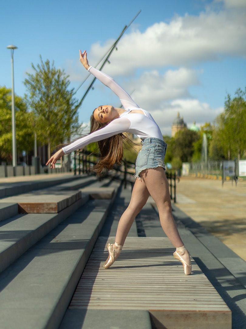 A ballerina in white bodysuit and denim shorts performs an elegant pose on pointe shoes outdoors.