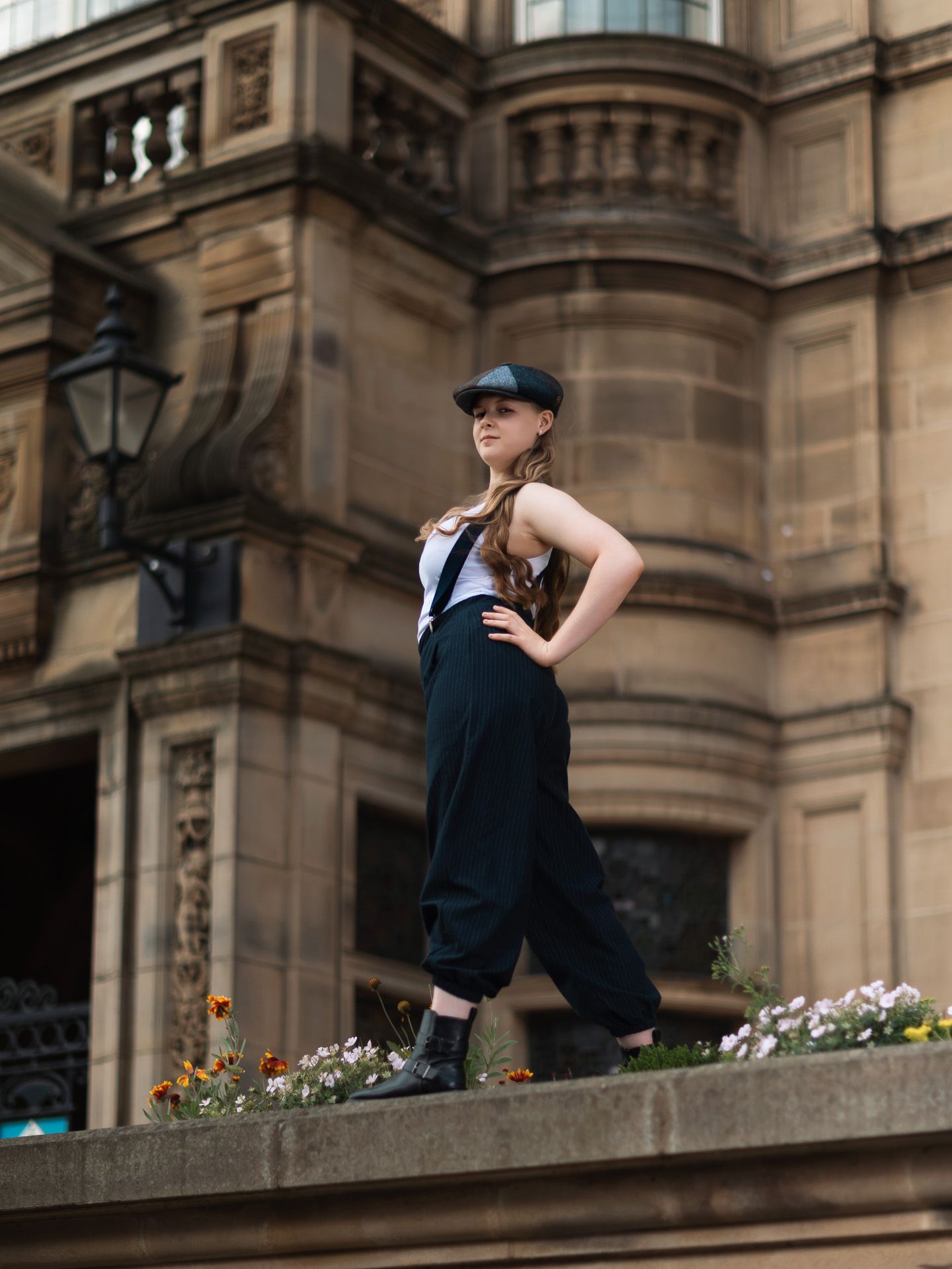 Young woman in black cap and suspenders poses on a stone ledge before a grand historic building with flowers.