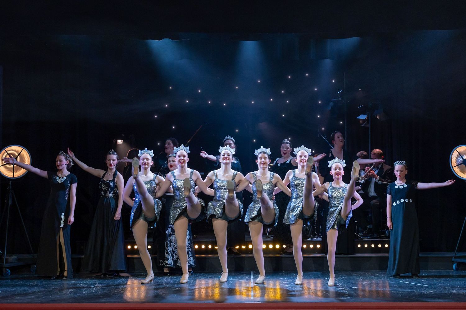 Chorus line dancers in silver costumes and headdresses perform on a theatrical stage with bright lights.