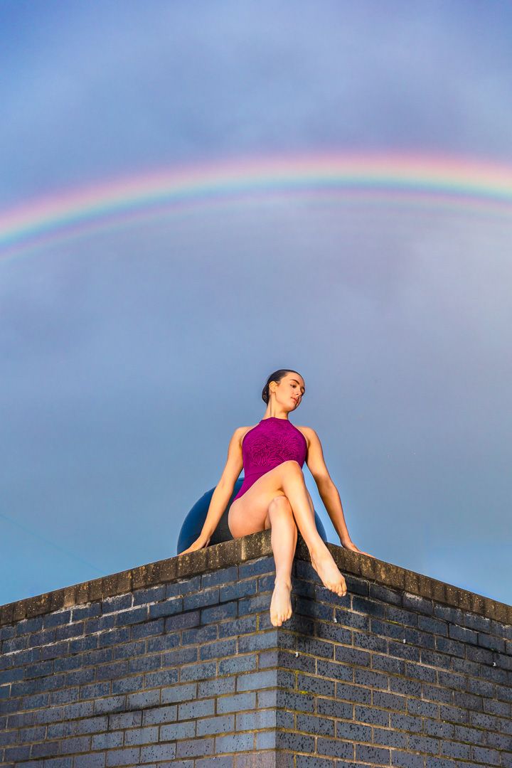 Dancer in purple leotard poses atop brick wall under vibrant rainbow in sky.