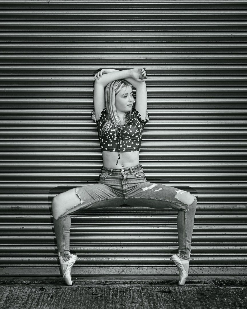 Black and white photo of a blonde dancer in ripped jeans and pointe shoes posing against a metal shutter.