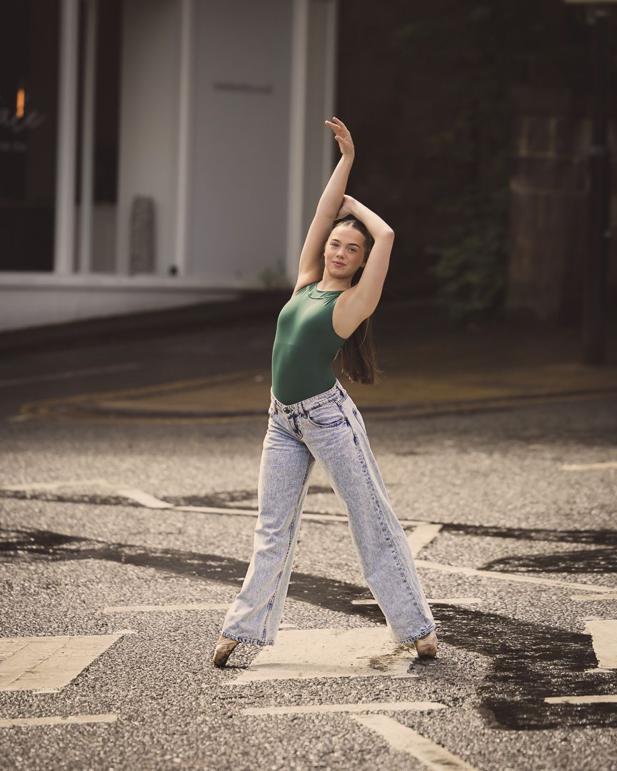 Young woman in green tank top and light wash jeans posing gracefully on a street crosswalk.