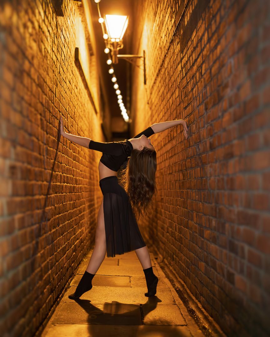 A figure in black dances through a dimly lit narrow brick alleyway illuminated by warm overhead lights.