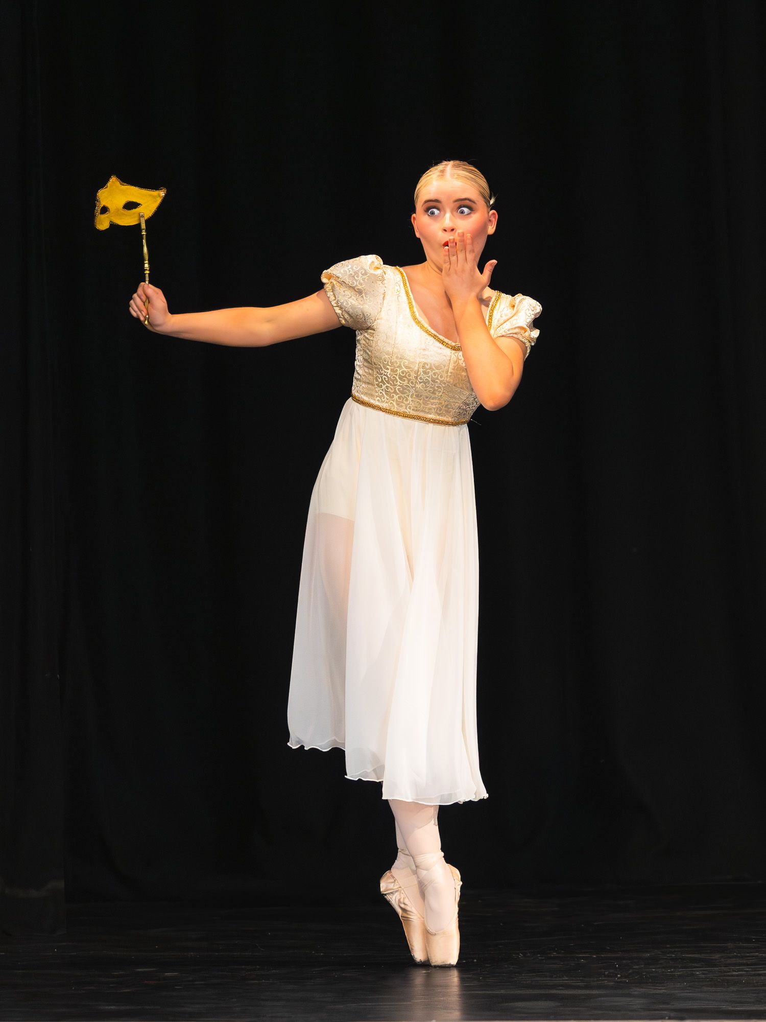 Ballet dancer in white dress holds yellow mask while performing en pointe.