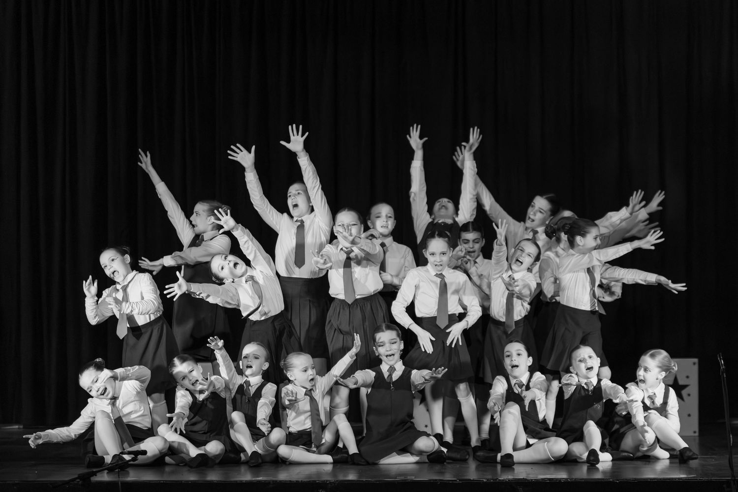 Black and white photo of children performing on stage, arms raised in a joyful dance recital finale.