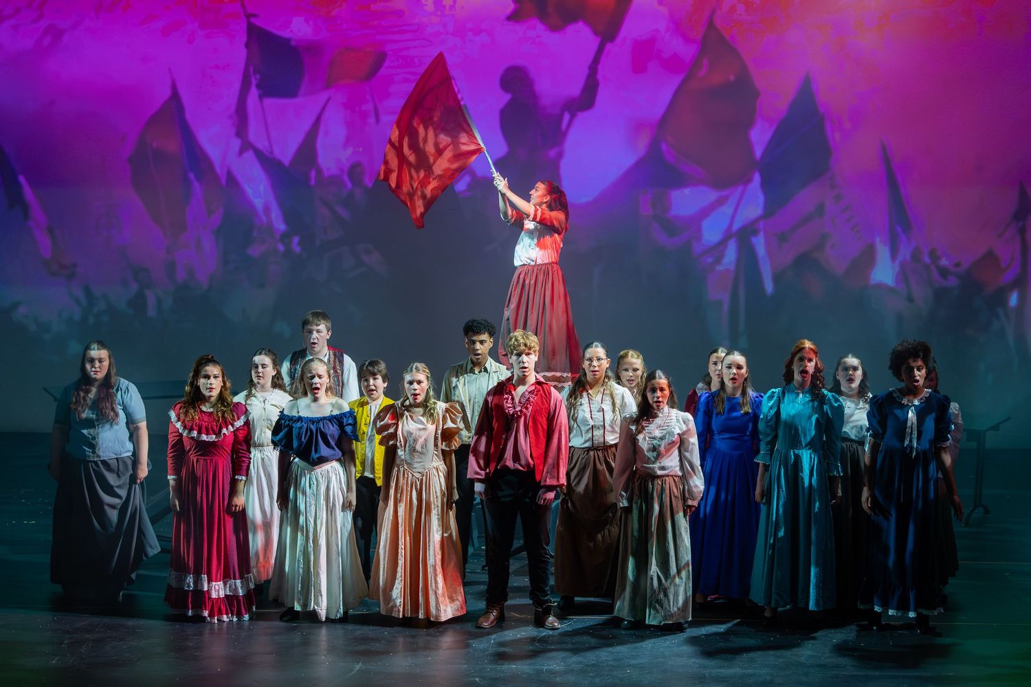 A theatrical ensemble in colorful costumes performs on stage, with a performer raising a red flag above the group.