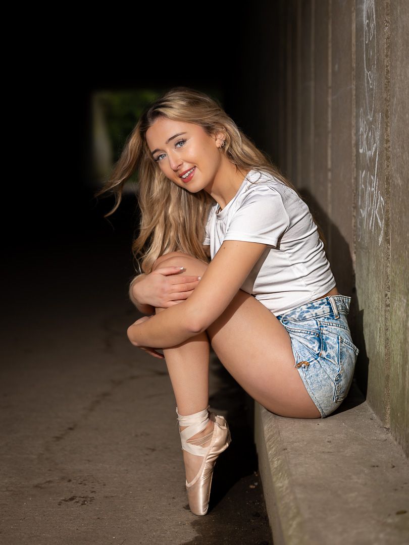 Dancer in white shirt and shorts poses gracefully against concrete wall with ballet pointe shoes.