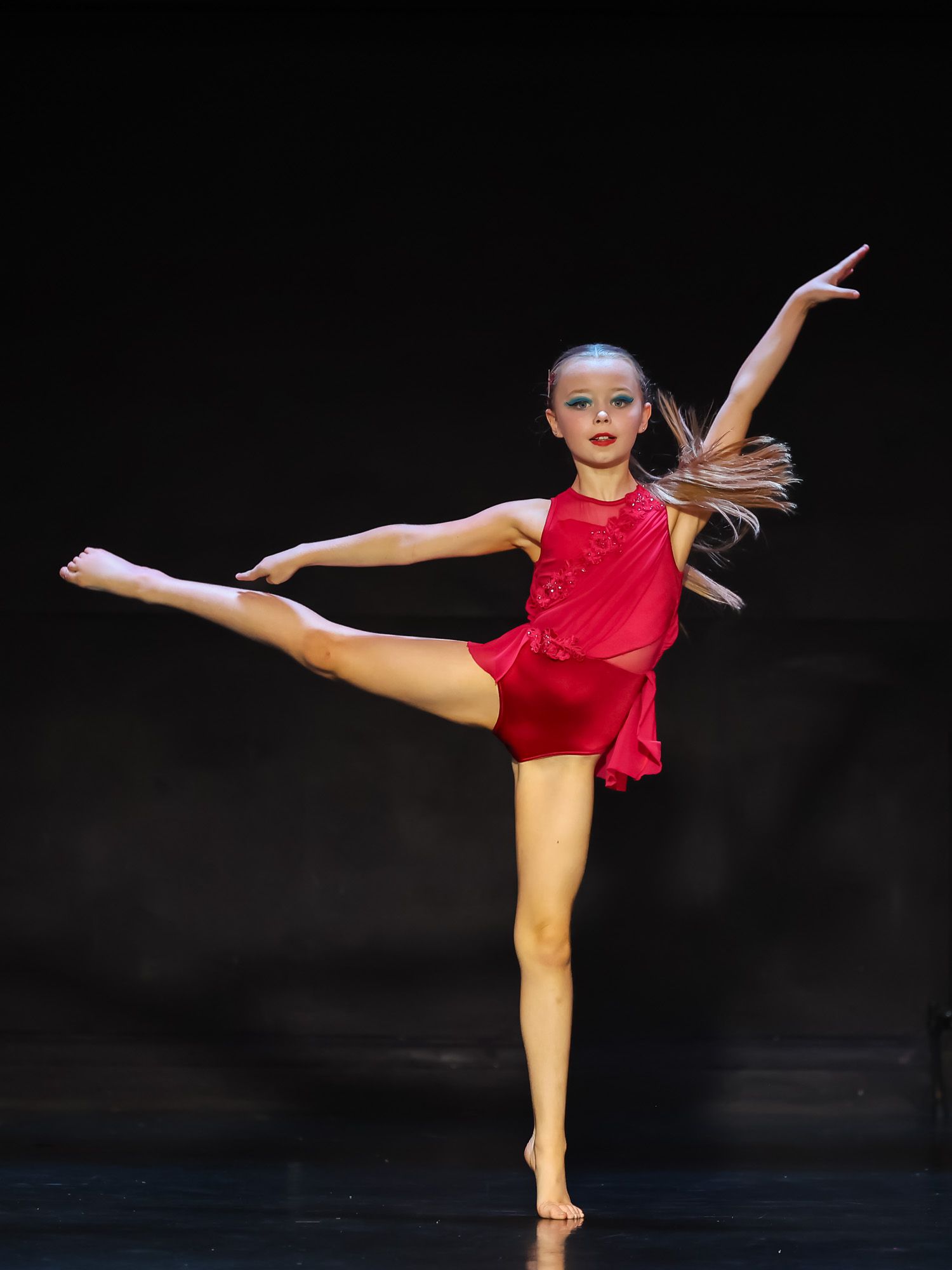 Young dancer in red costume performs an elegant arabesque on stage under spotlight.