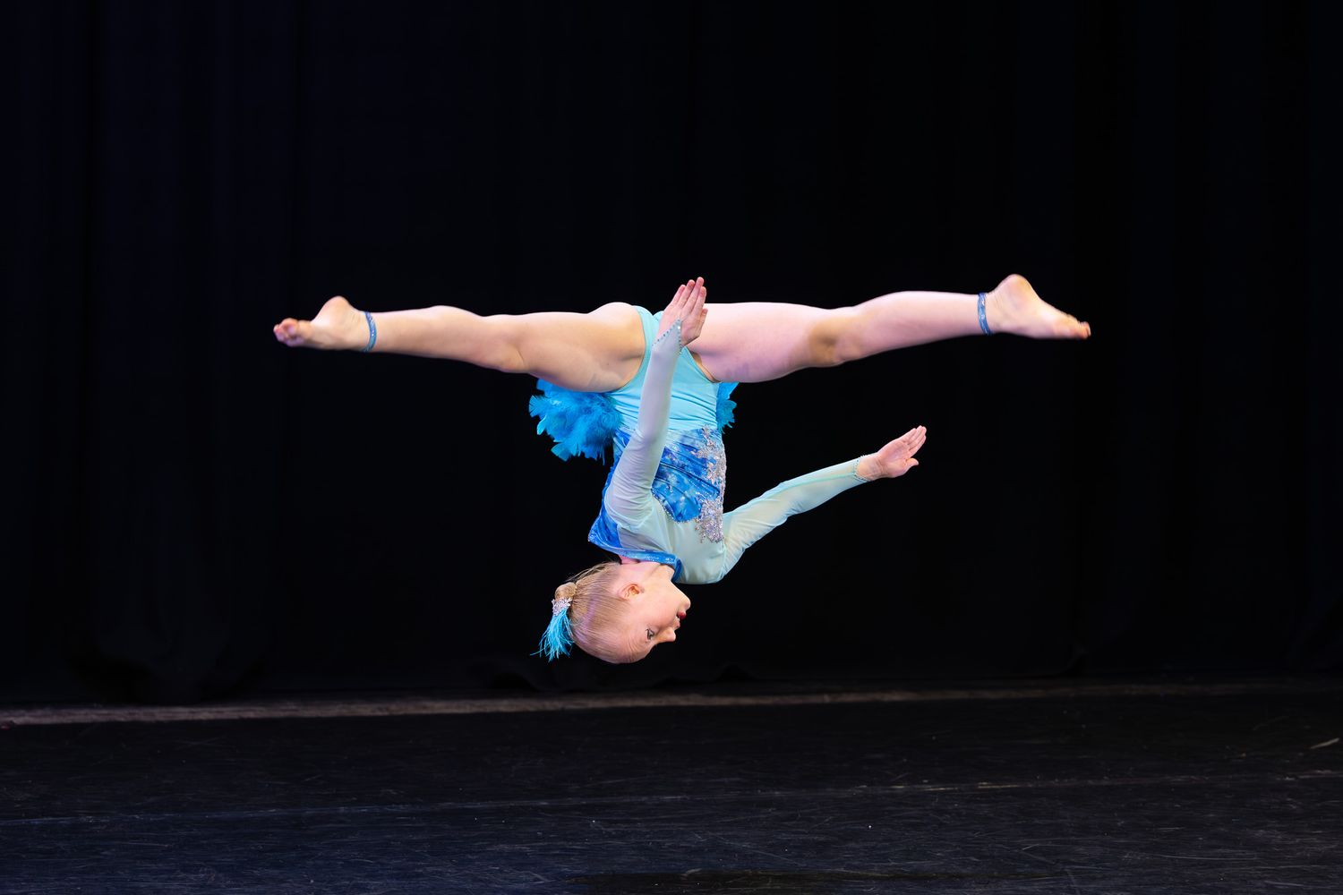 Young gymnast in blue costume performs a stunning back walkover on stage during a dance recital.