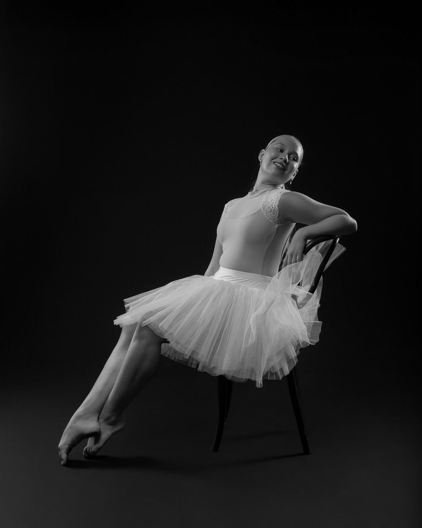 Black and white photo of a young ballerina in white tutu sitting gracefully on a chair in a dance studio.