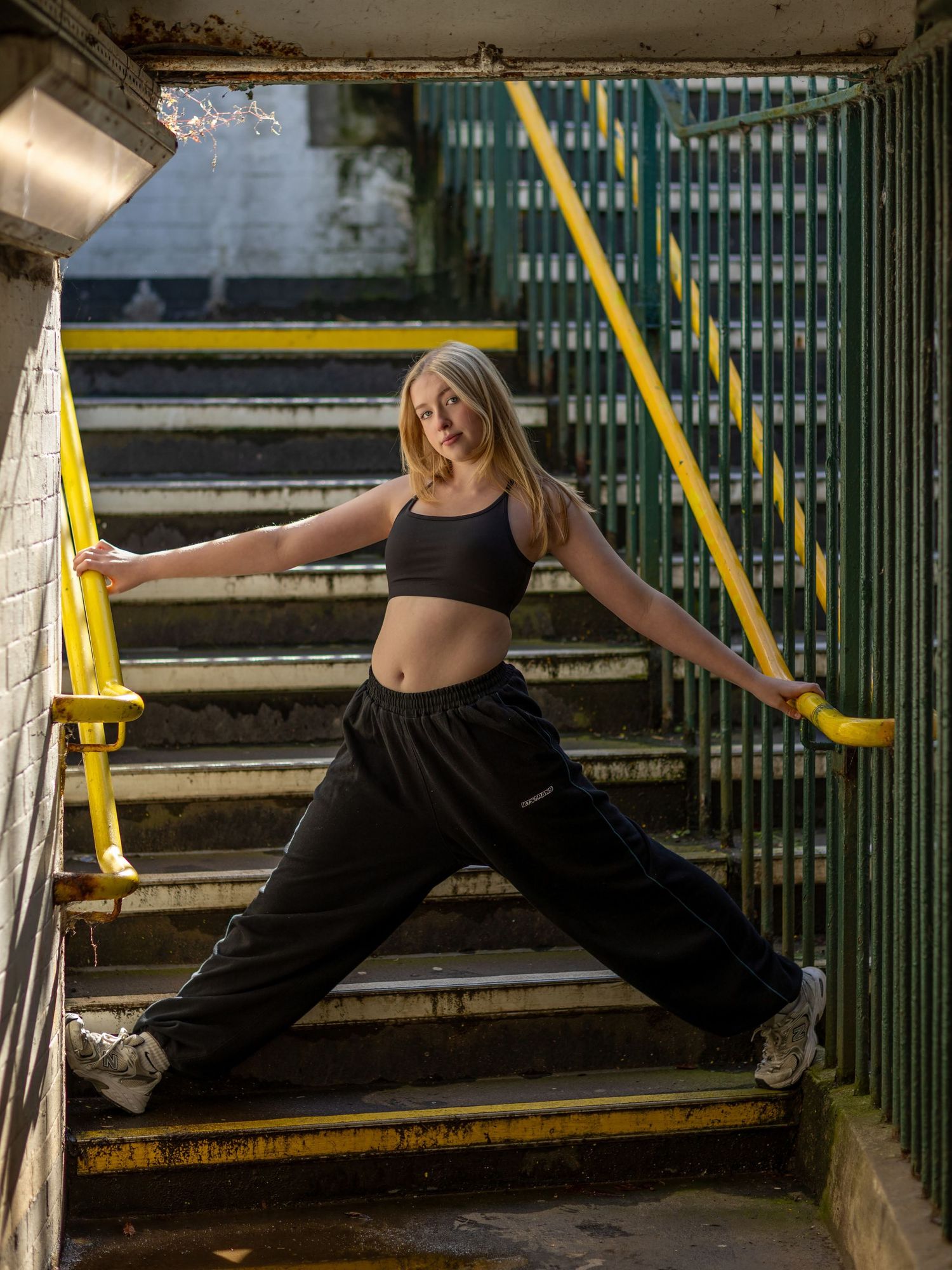A person in black athletic wear performs a split stretch across concrete steps in a dimly lit urban setting.