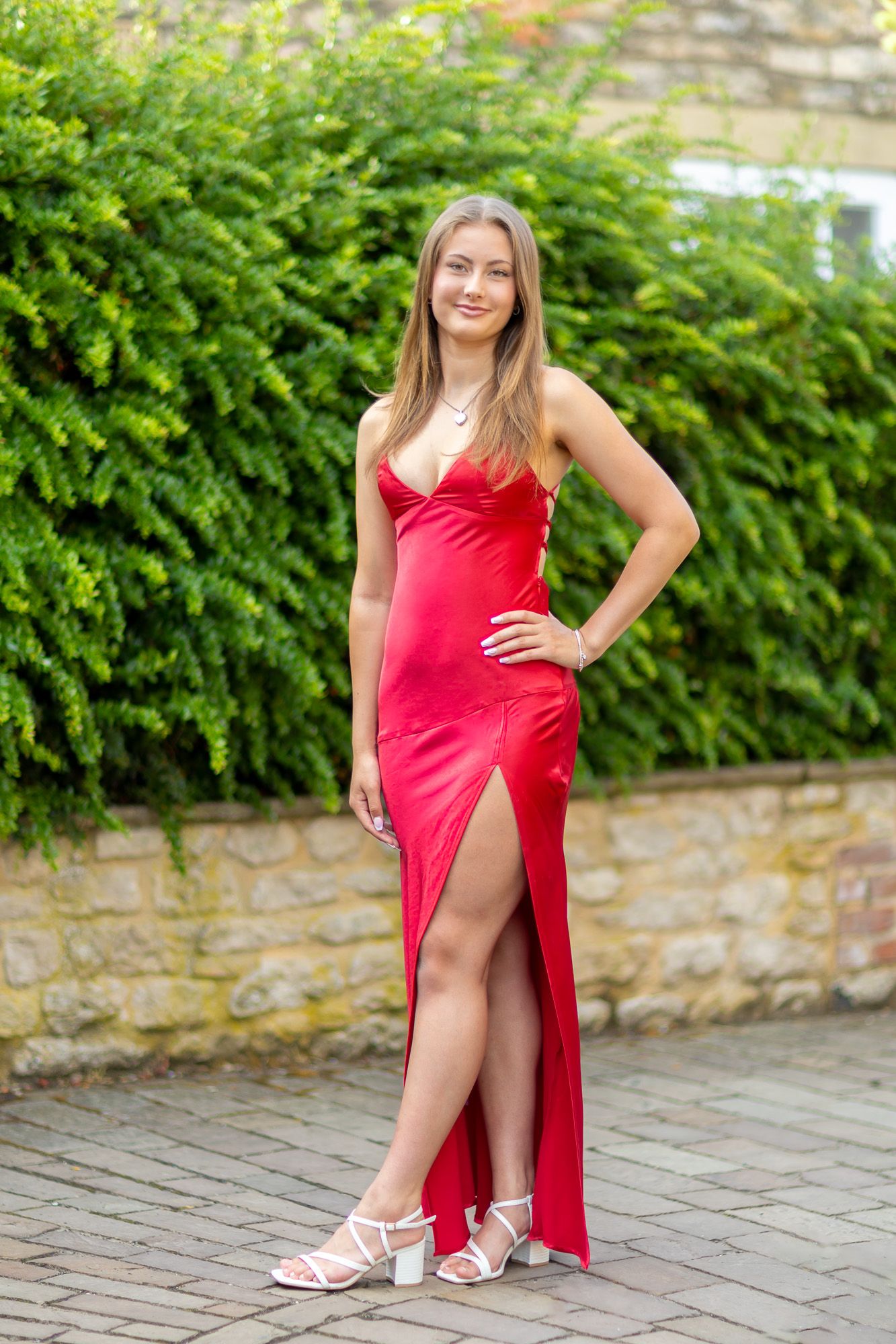 A person in a vibrant red formal dress poses against a stone wall and greenery.