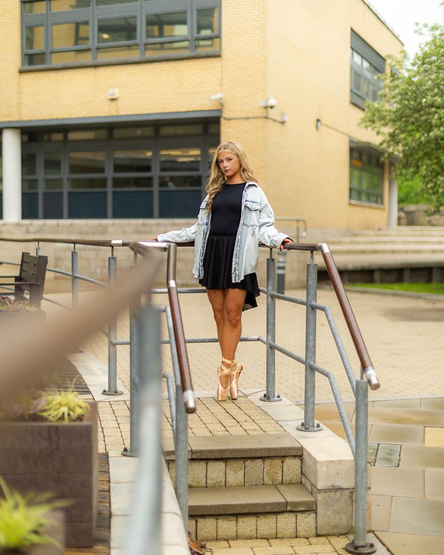 Young woman in black dress and denim jacket poses on steps wearing ballet pointe shoes outside a brick building.
