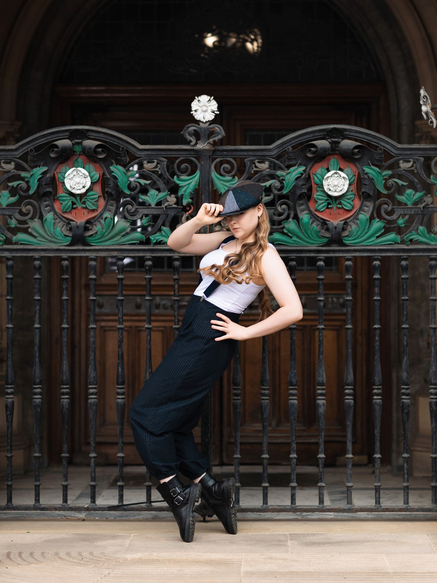 Young woman in black hat, suspenders, and boots poses dynamically before ornate iron gates.