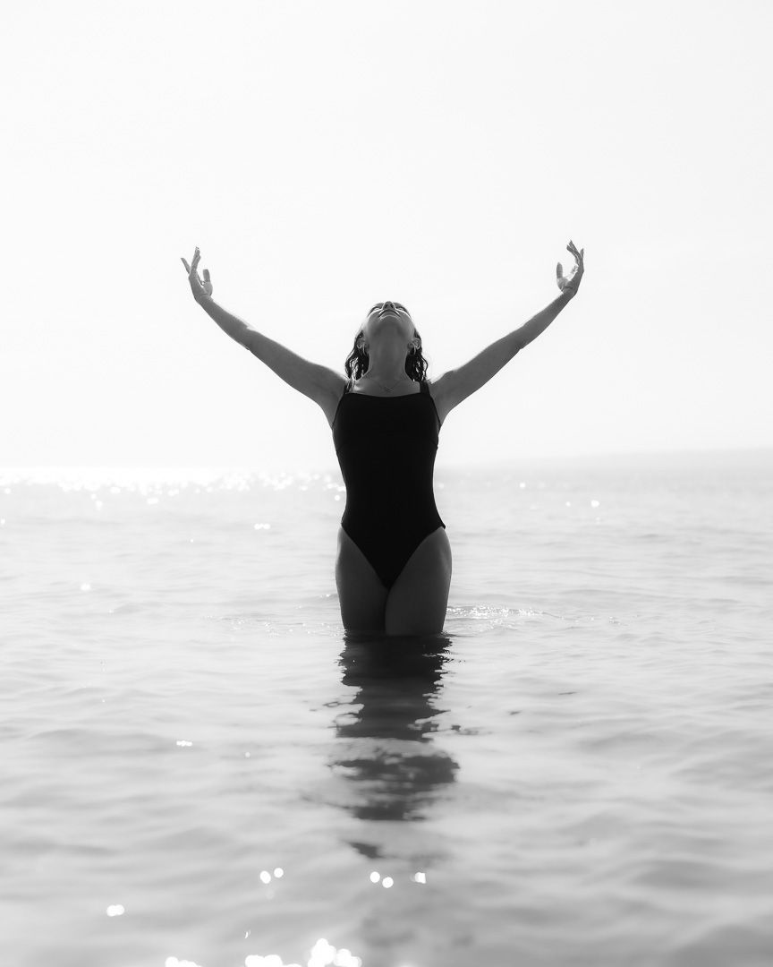 Woman in black swimsuit standing in ocean with arms raised in black and white photo.