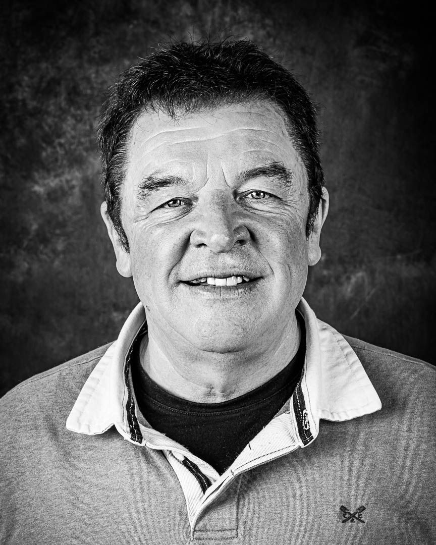 Black and white portrait of a smiling middle-aged man wearing a polo shirt against a textured dark background.