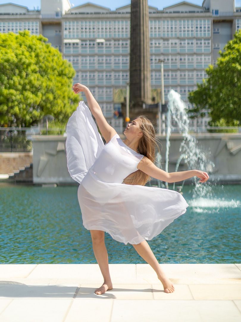 A woman in a flowing white dress dances gracefully near a fountain with a large building in the background.