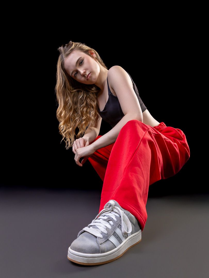 Young woman in red pants, black crop top, and gray sneakers poses seated on studio floor.