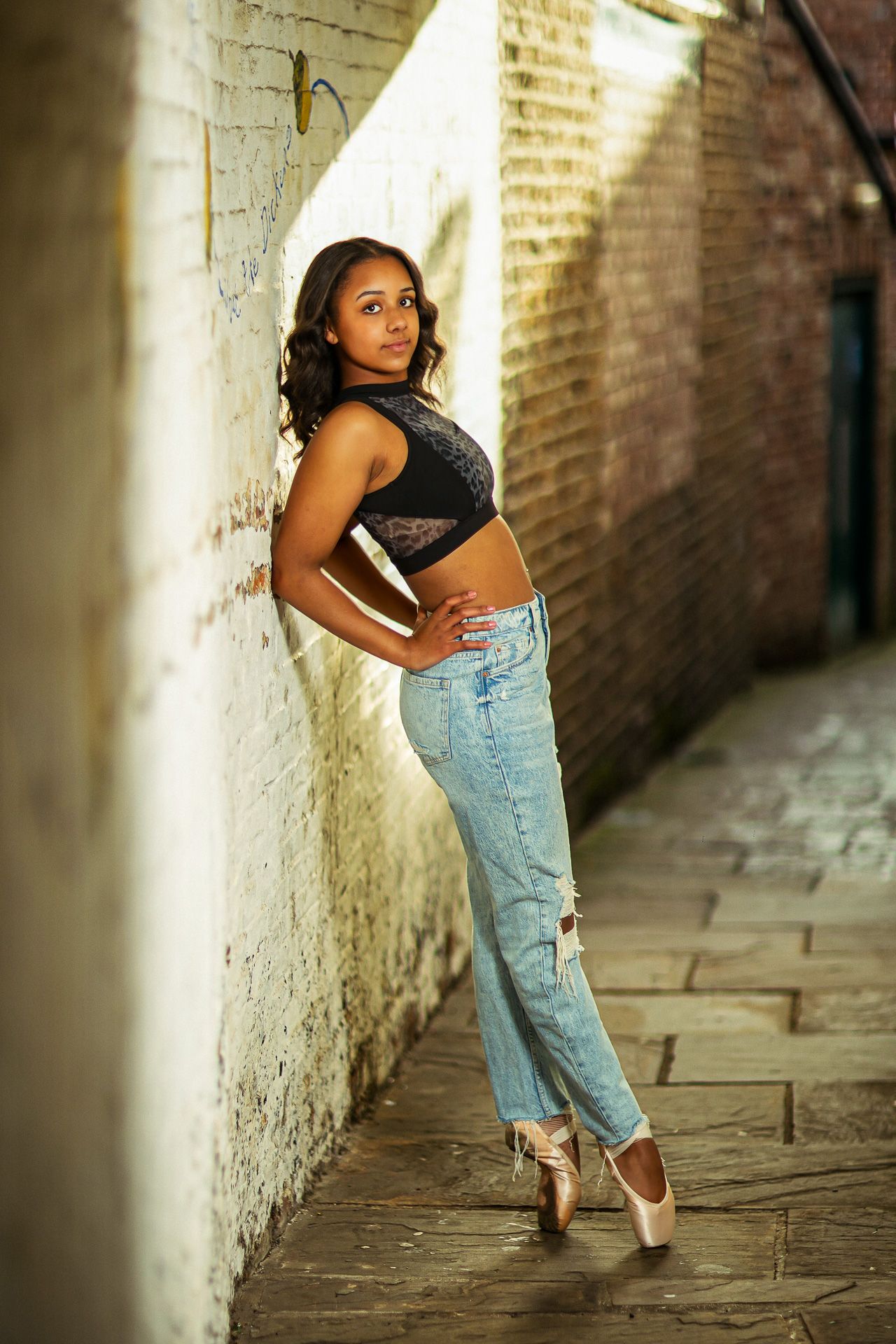 A young Black ballerina in jeans and a crop top poses en pointe in a narrow brick alleyway.