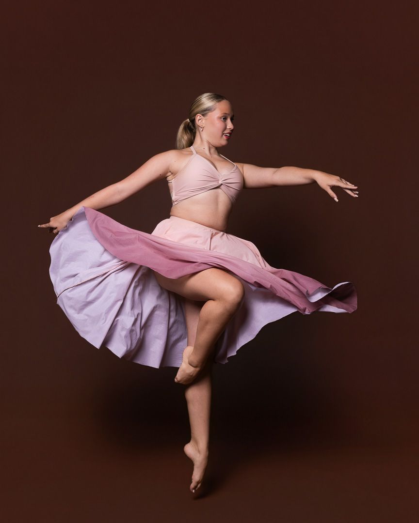 A blonde female dancer in a pink crop top and flowing skirt performs an elegant pirouette on a brown background.