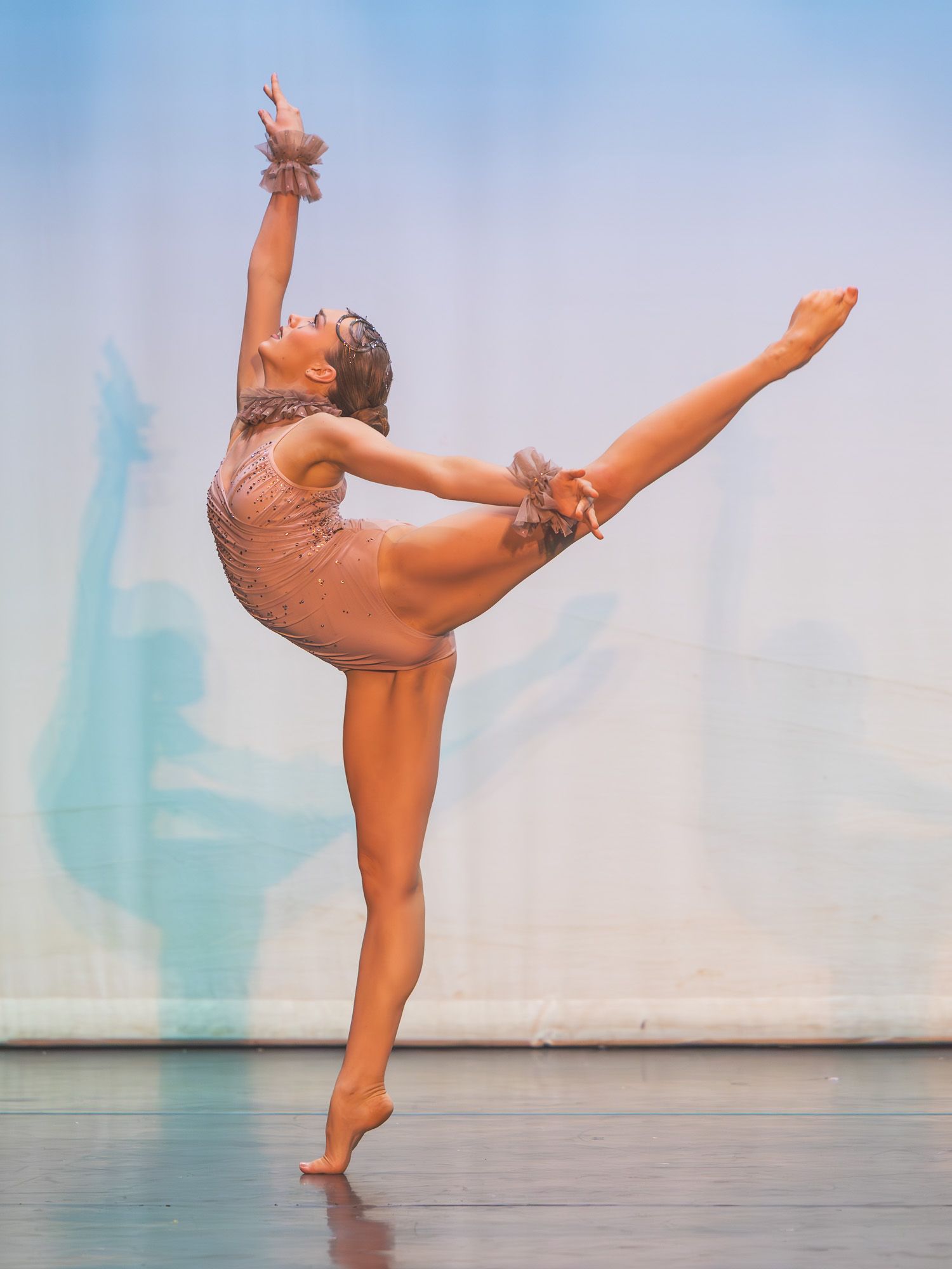 A dancer performs an arabesque on stage in a sparkly nude costume under blue stage lighting.