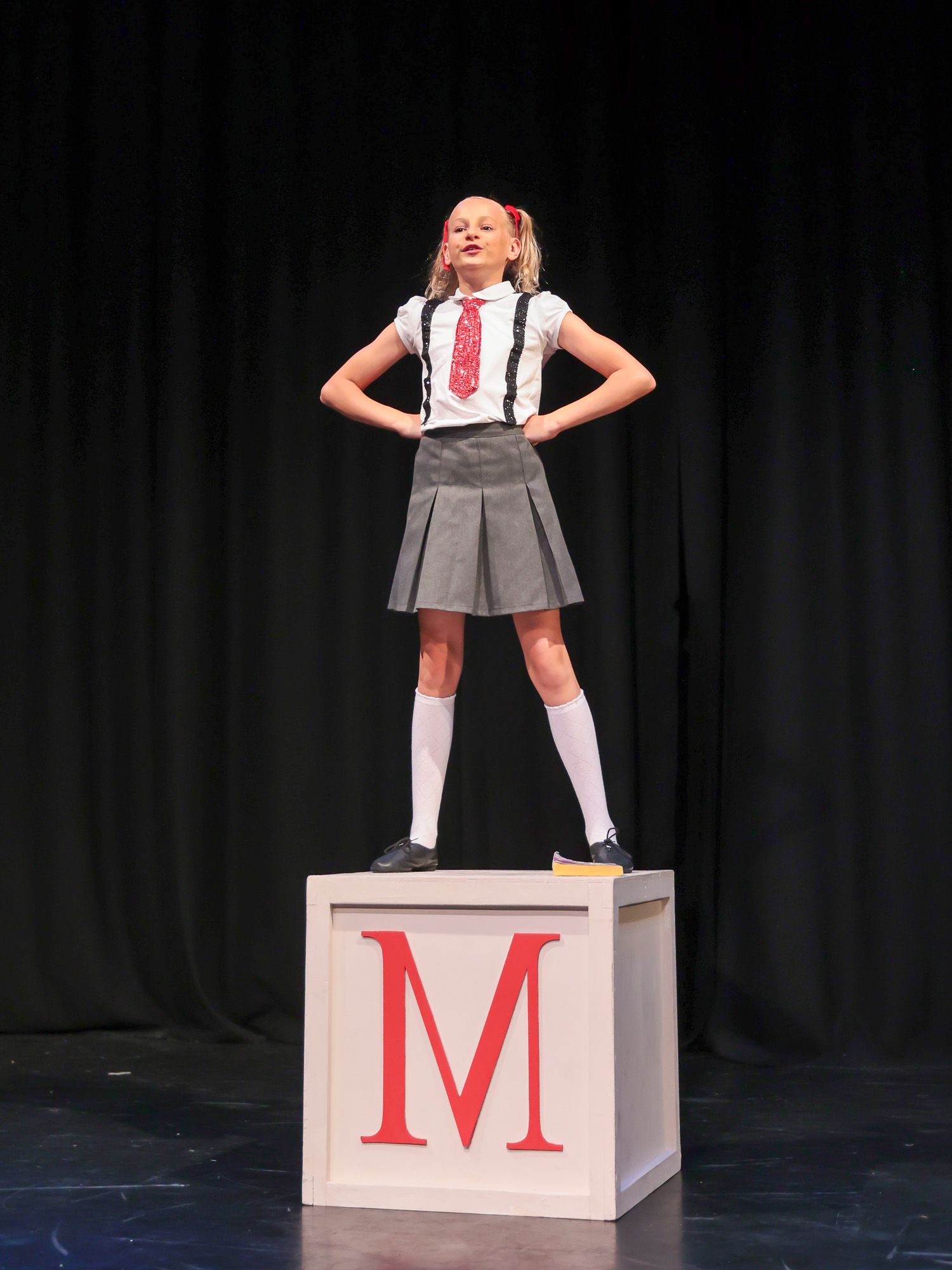 Young girl in school uniform stands confidently on a large wooden block with red letter M on a stage.