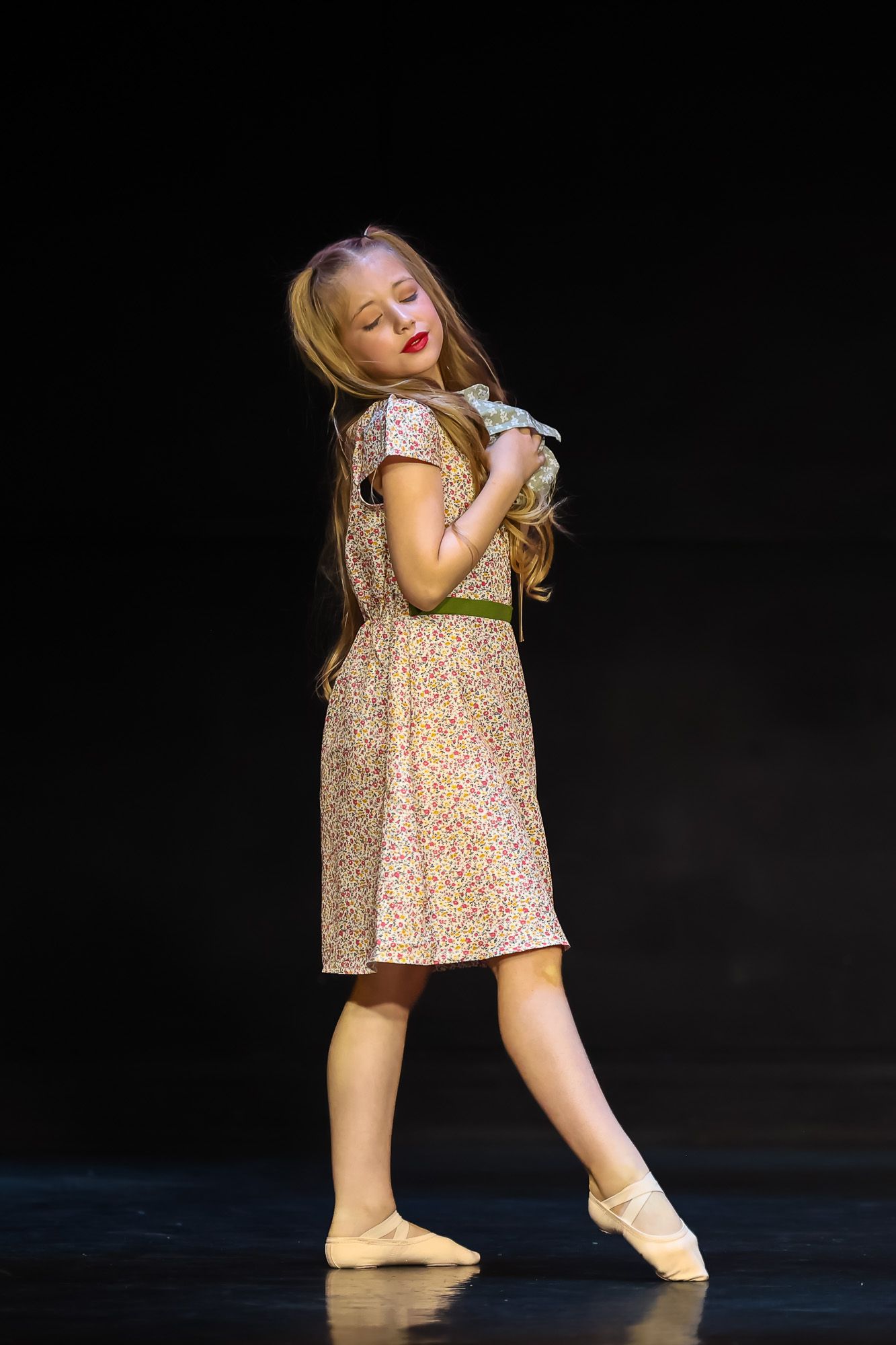 Young girl in floral dress and ballet slippers performs graceful dance pose on dark stage.