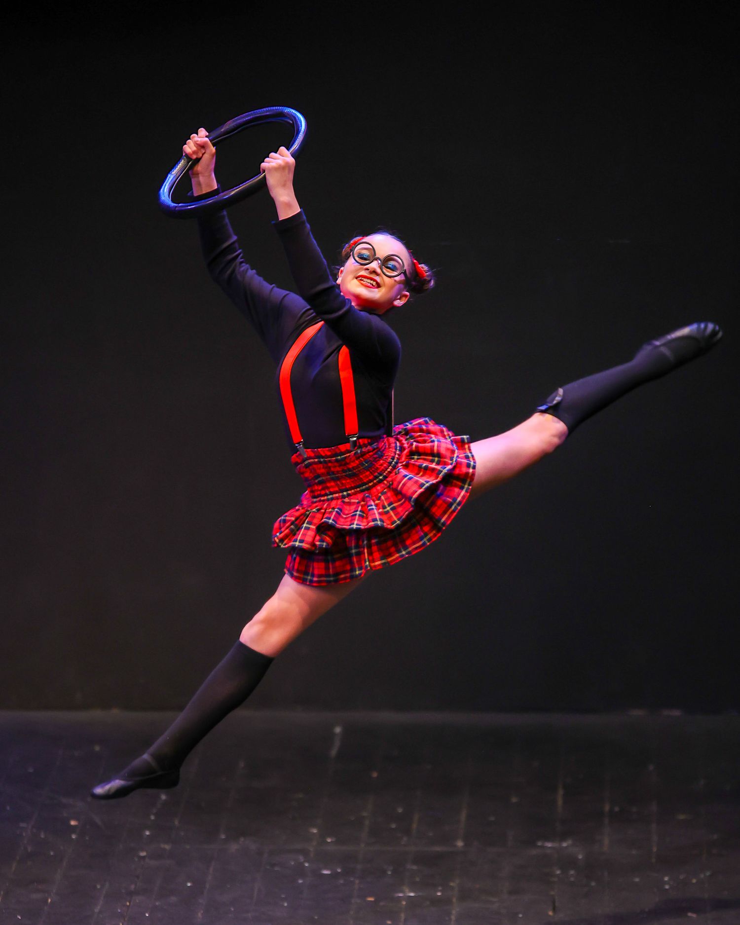 Young dancer leaps gracefully in plaid skirt holding blue hoop, wearing glasses and black costume on stage.