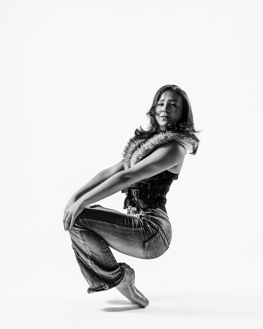 Black and white photo of a dancer in jeans and fur vest, striking a dynamic crouching pose against white background.