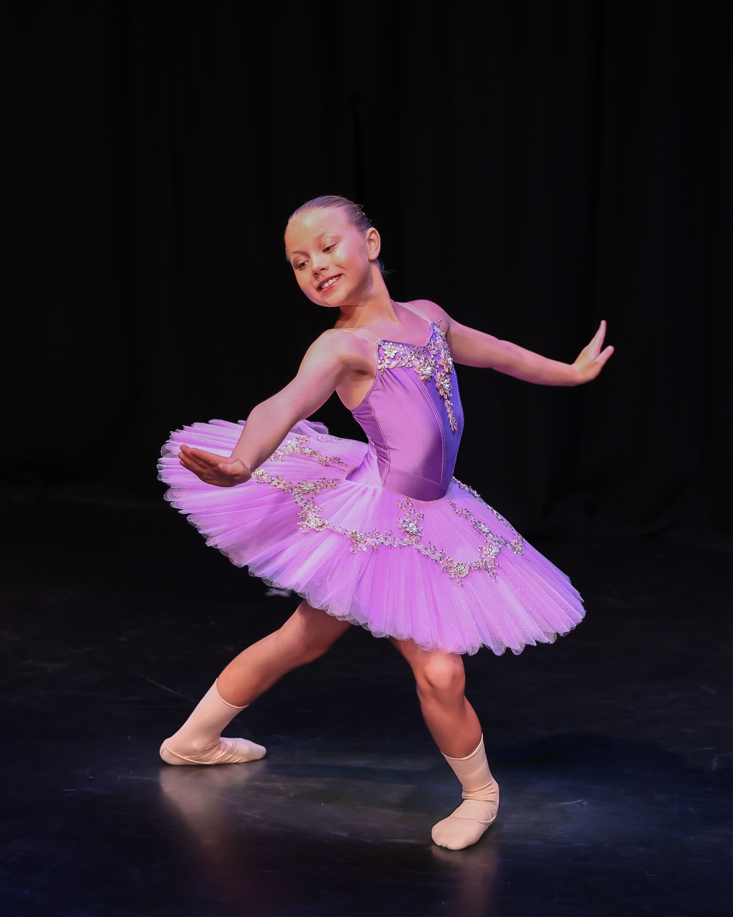 Young girl performing ballet on stage in a purple tutu costume with jeweled embellishments under bright spotlights.