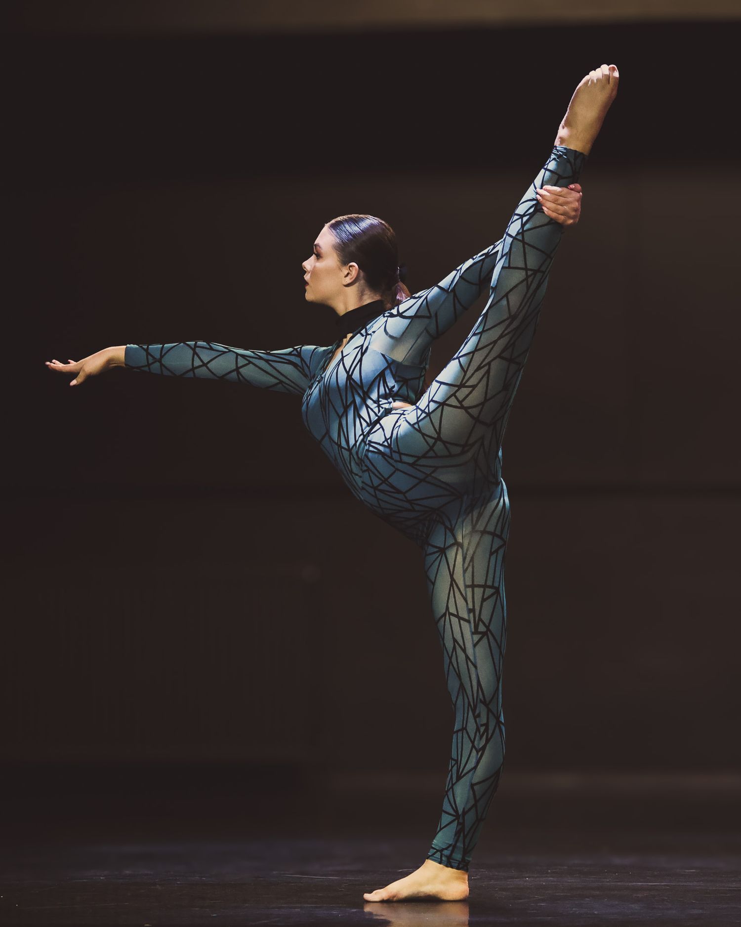 A dancer in a patterned teal bodysuit executes a high leg extension on stage during a contemporary performance.