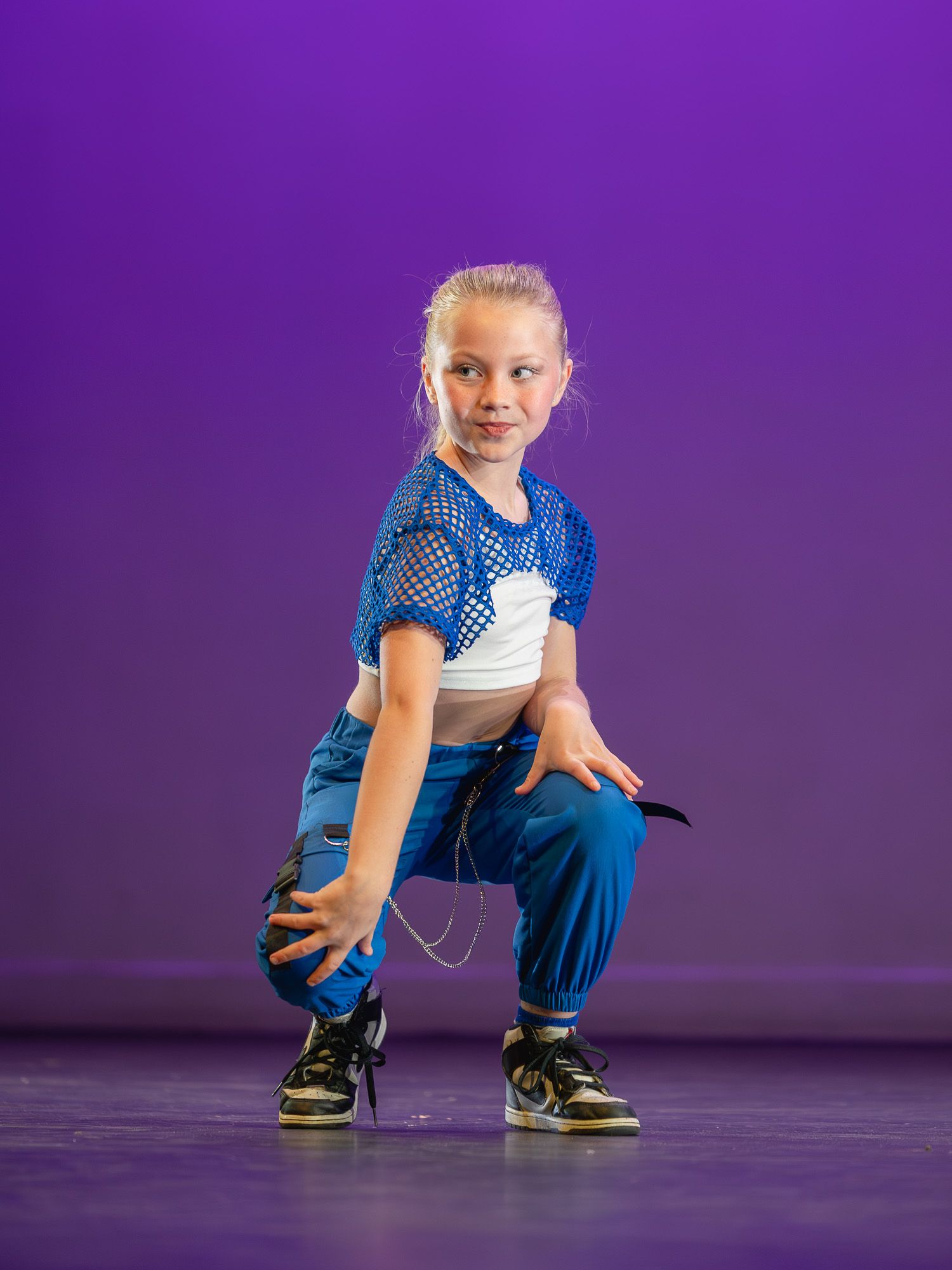 Young girl in blue mesh top and teal pants strikes a hip-hop dance pose on a purple-lit stage.