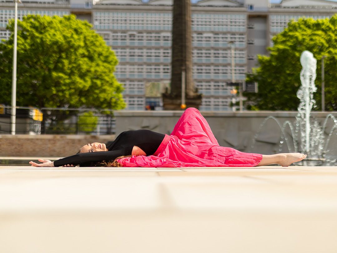 A dancer in a red skirt and black top lies gracefully on a sunny plaza with fountains and trees in the background.