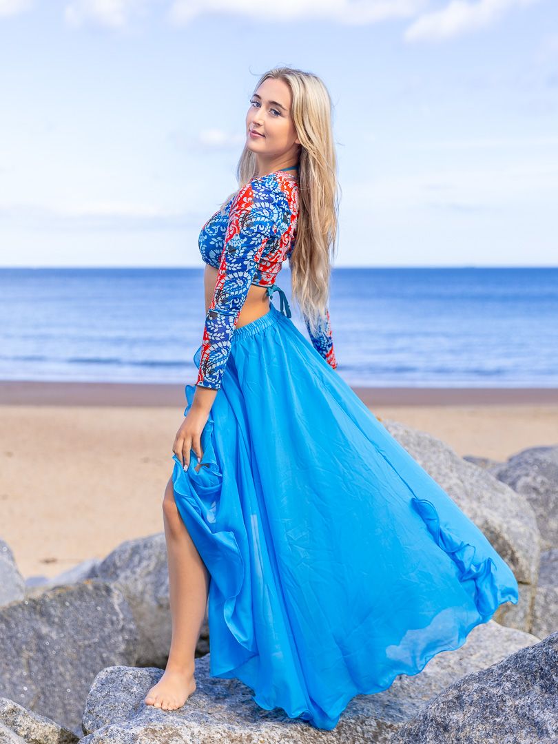 Blonde woman in blue maxi skirt and patterned top posing on rocks at a sandy beach with ocean backdrop.